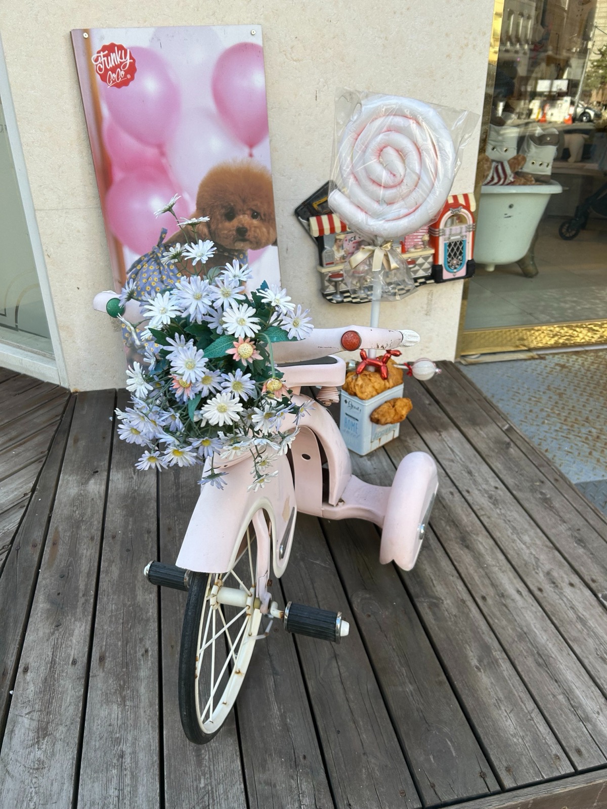 ペットショップの店先。花を飾ったピンクの三輪車、キャンディ形の犬用おもちゃ、トイプードルのポスターが置かれている